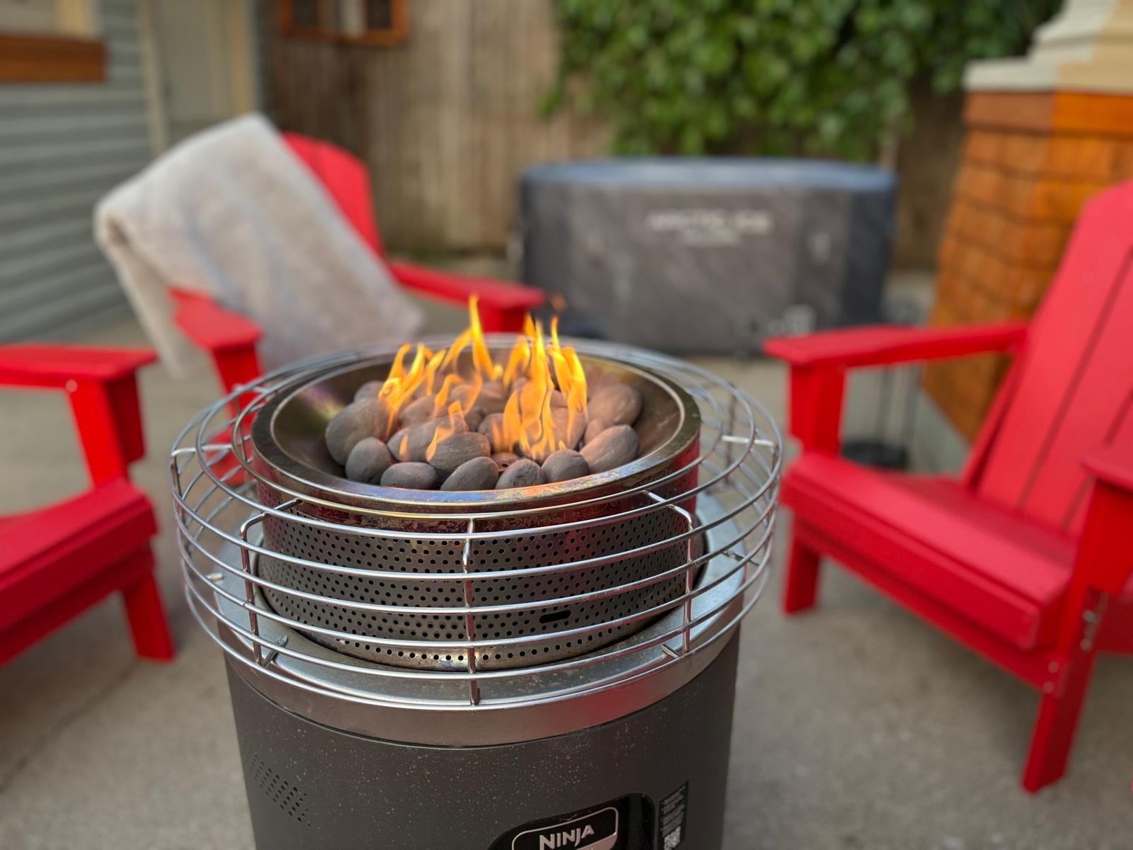 Propane fire pit with flames, red Adirondack chairs and cold plunge tub in the background